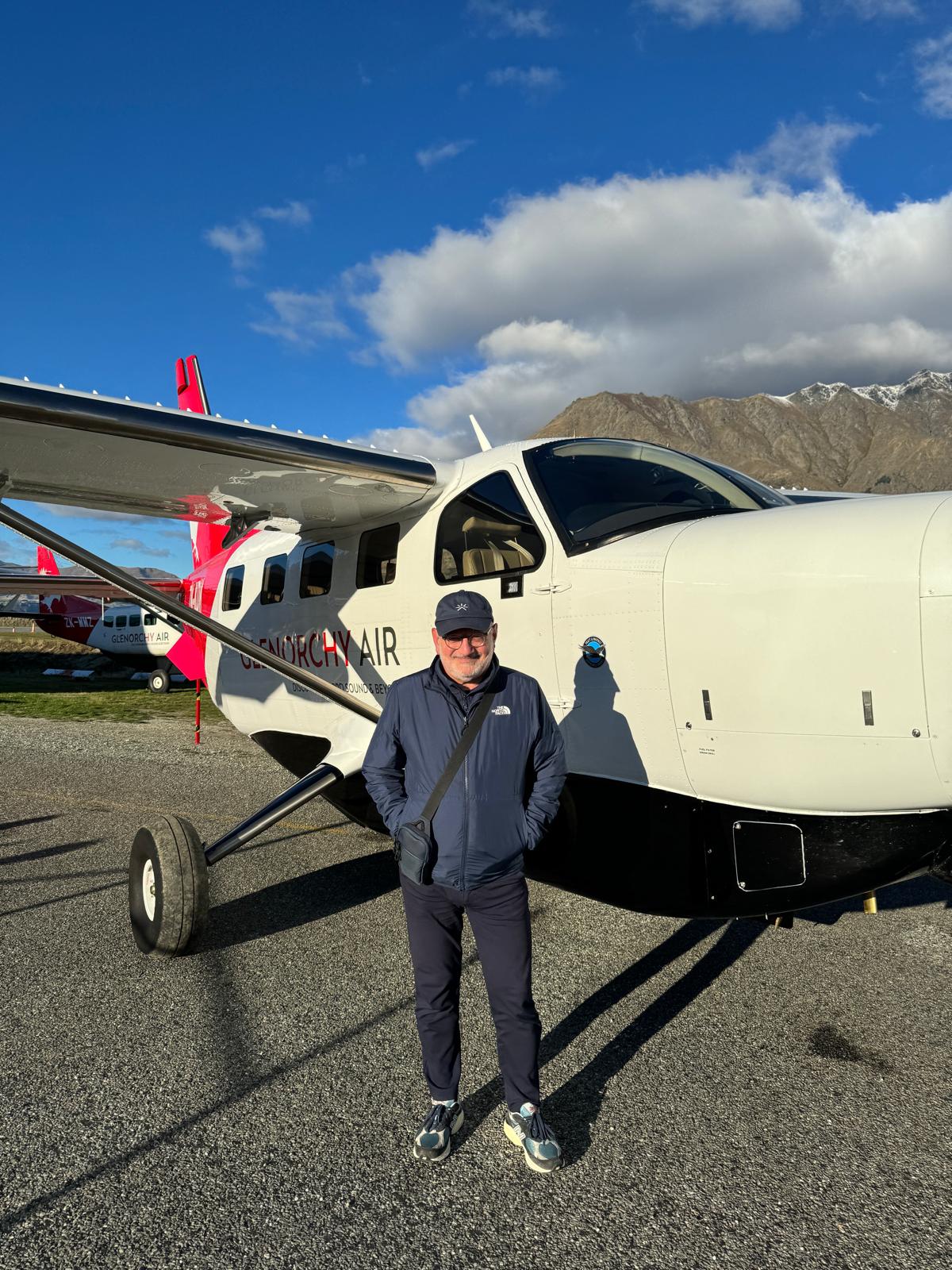 George Jerjian standing in front of a small white aircraft with 'Glentorchy Air' branding, set against a stunning mountain backdrop with a bright blue sky. He is dressed in a navy jacket, dark pants, and a crossbody bag, smiling and ready for adventure.